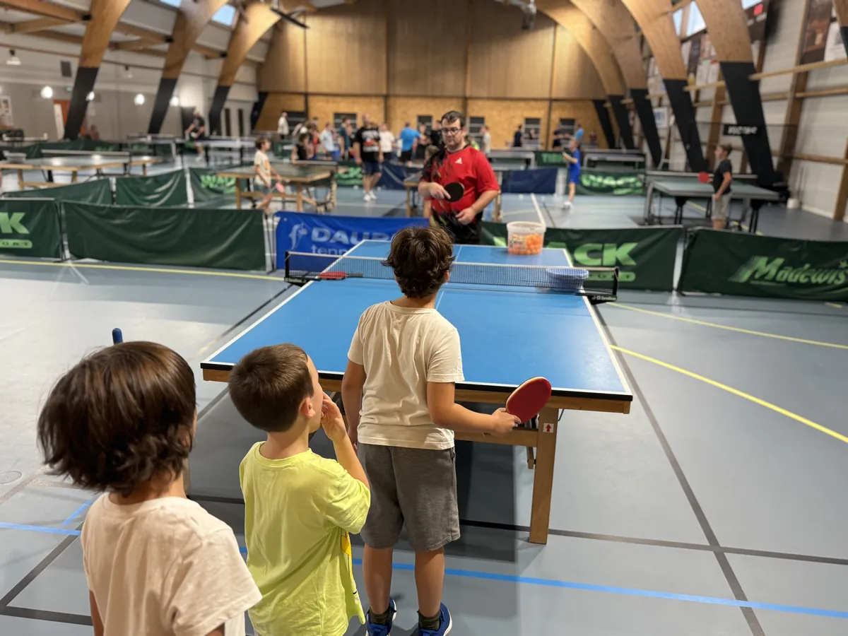 Trois enfants de dos, debout devant une table bleue, regardent un entraîneur en maillot rouge qui tient sa raquette, dans la grande salle.