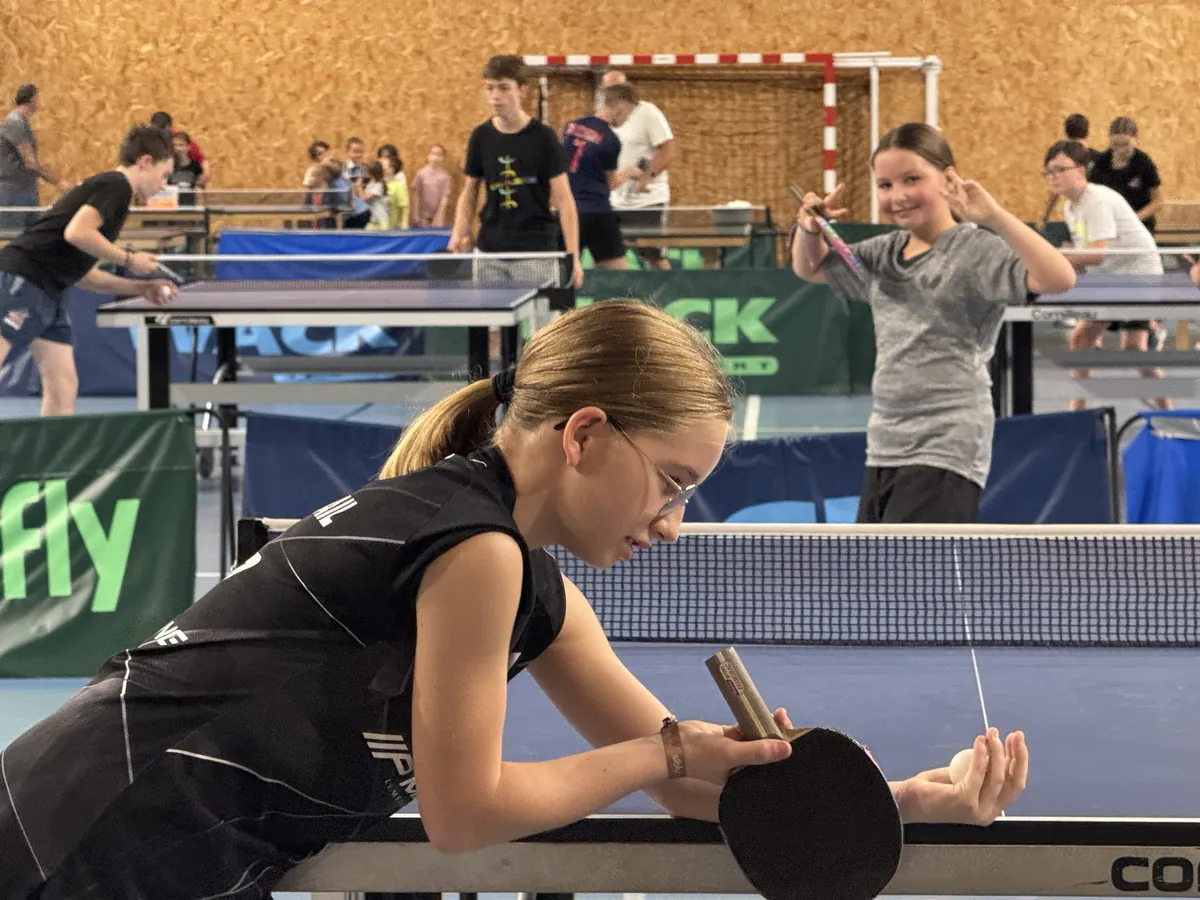 Zoé, jeune joueuse concentrée, la balle dans la main et la raquette posée sur la table, s'apprête à servir ; une autre sourit derrière elle.