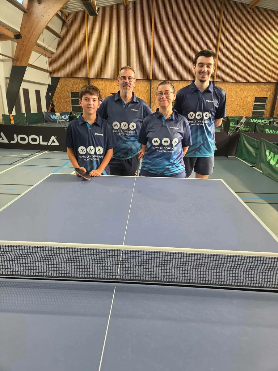 Un garçon, deux hommes et une femme en maillot du club, debout derrière une table de tennis de table dans une grande salle.