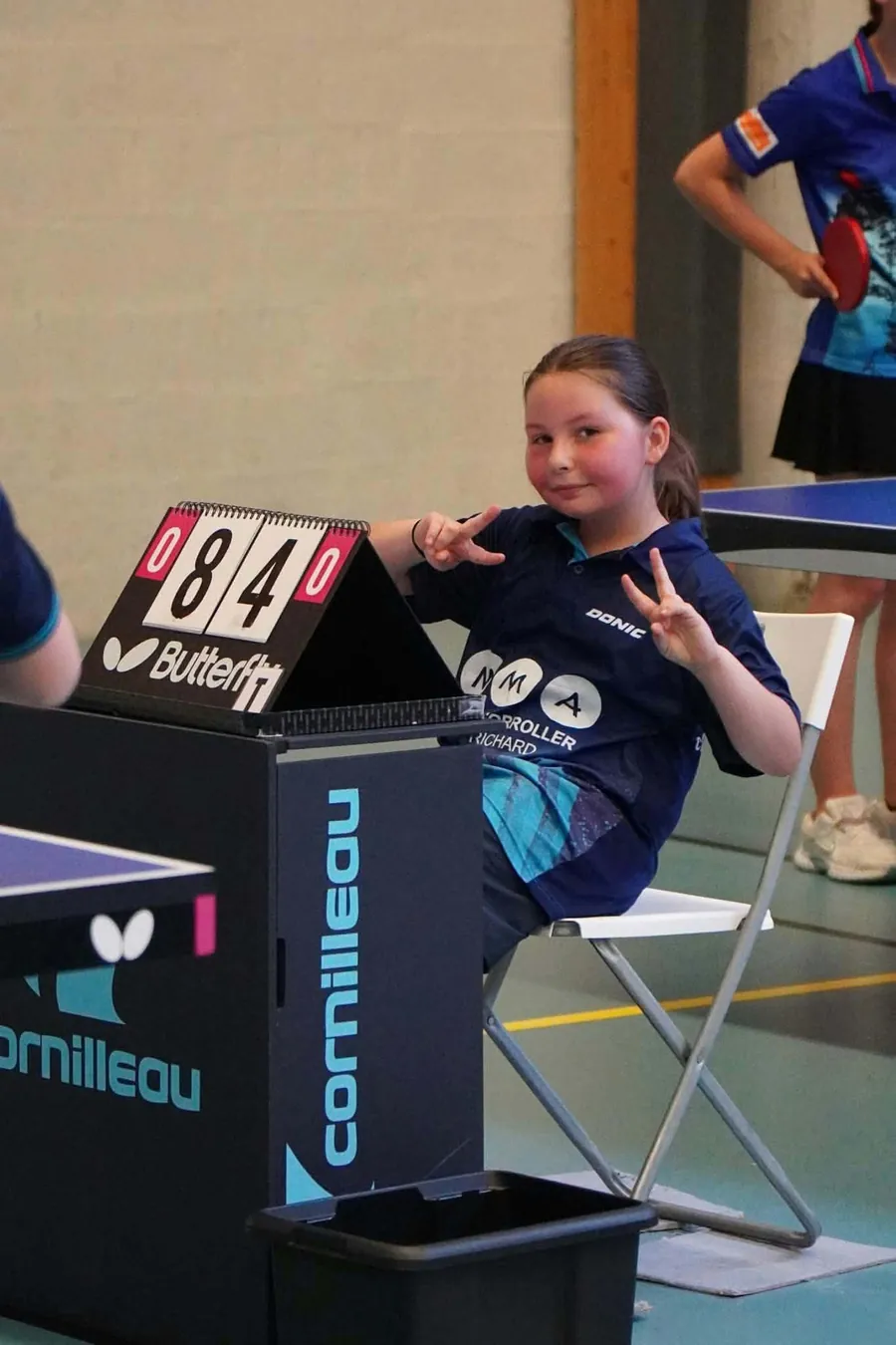Une jeune joueuse en maillot du club, assise à côté du marqueur de points, fait le V de la victoire des deux mains.